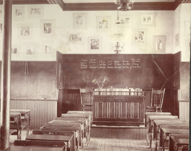 Interior view of the PLeasant Hill school. There are neat rows of school desks and at the front, a large desk for the teacher. Seventeen pictures from about 1900 are hung on the walls above the blackboards.